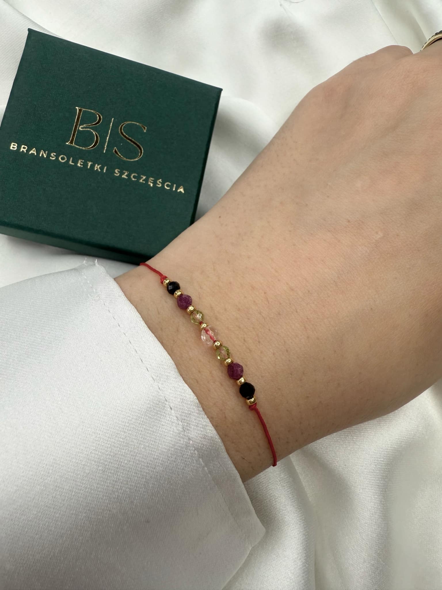 Zbliżenie na bransoletkę na czerwonym sznurku z naturalnymi kamieniami (kryształ górski, oliwin, rubin, czarny turmalin), na nadgarstku, na białym tle.