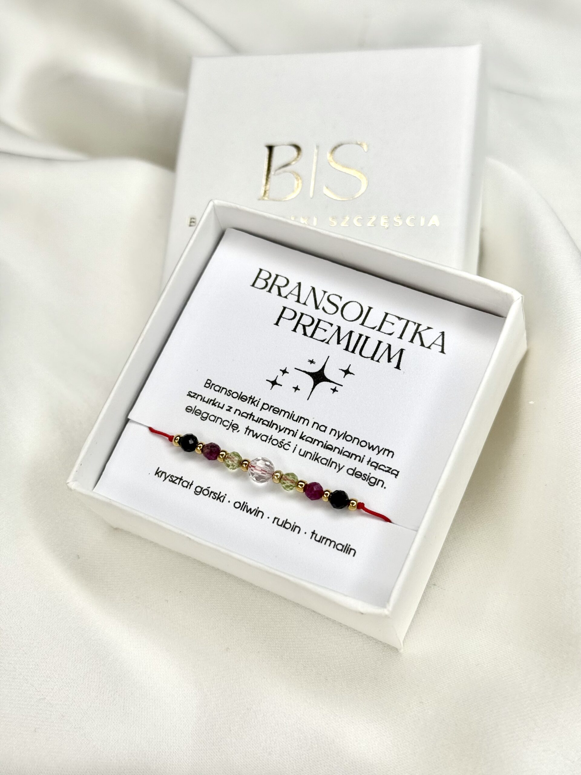 Bransoletka na czerwonym sznurku z naturalnymi kamieniami: kryształ górski, oliwin, rubin i czarny turmalin, na nadgarstku, na białym tle.