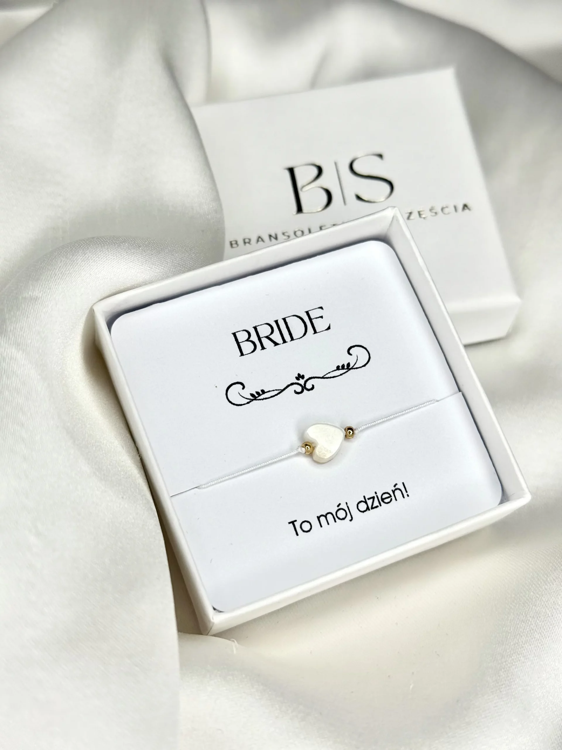 Bransoletka BRIDE z serduszkiem z masy perłowej i złotymi koralikami na białym sznurku, prezentowana w pudełku.